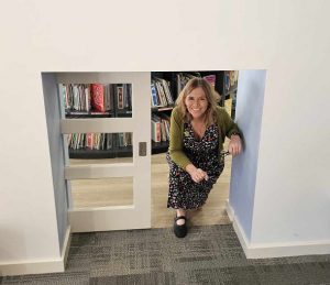 author Sally Murphy is on her knees coming through a child sized door in a library. 
